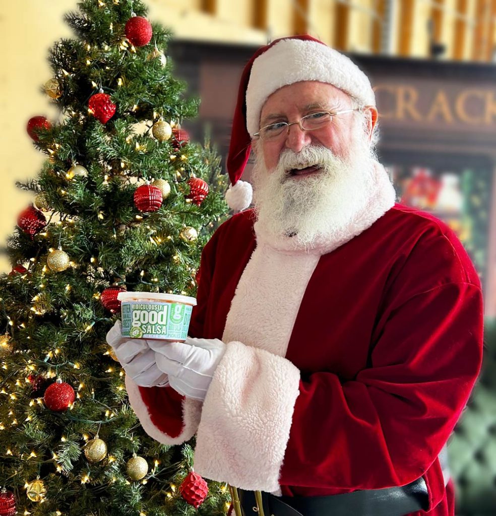 Santa holding Ridiculously Good Salsa beside Christmas tree